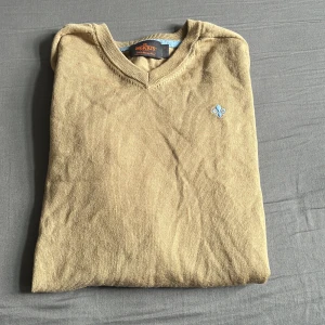 Beige merinoullströja från Morris - Snygg beige tröja från Morris i 100% merinoull med v-ringning och diskret ljusblå broderad logga på bröstet. Långärmad modell med ribbade muddar och klassisk passform. Perfekt för dig som gillar stilrena och bekväma plagg. Hör gärna av er vid minst lilla fråga 