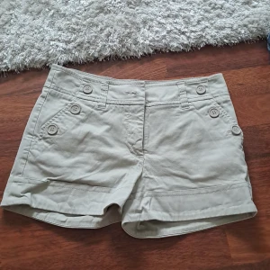 Beige shorts med knappar från H&M - Snygga beige shorts från H&M med låg midja och dekorativa knappar framtill. Klassisk modell med fickor på sidorna och bakfickor. Tillverkade i bomull, perfekta för varma dagar. Personligen tycker jag att byxorna är coola och unika!😍 Säljer för att dom tyvärr är för små. Sparsamt använda.🥹💞 Passar nog 32 och 34. Jag brukar bära 36.