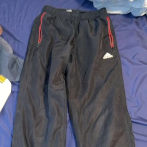 Svarta Adidas mjukisbyxor med röda detaljer - Svarta mjukisbyxor från Adidas med röda dragkedjor vid fickorna och klassisk Adidas-logga på låret. Byxorna har resår i midjan och är perfekta för träning eller chill. Snygg och sportig look med bekväm passform.