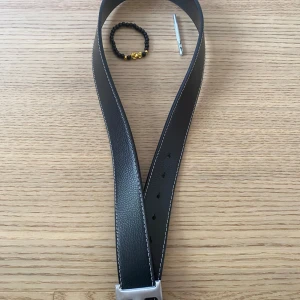Komprivat för fler bilder - Stilrent svart bälte i skinn med silverfärgat metallspänne. Bältet har vit kontrastsöm längs kanterna och klassisk design som passar till jeans eller kostymbyxor. Perfekt accessoar för att lyfta din outfit.