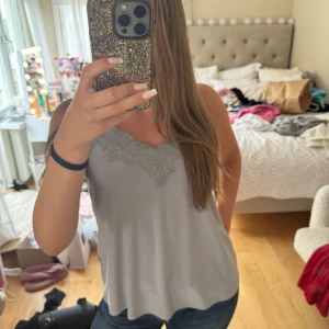 Ljusgråt linne med spetsdetaljer - Supersöt ljusgråt linne med tunna axelband och v-ringning dekorerad med spets. Toppen har en flowy passform och är perfekt att styla med jeans eller kjol. Storlek M men sitter även jättefint på folk som har storlek S. Spetsen ger en extra touch och gör toppen både trendig och feminin!