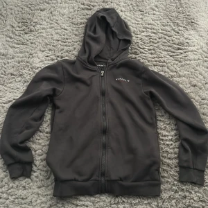 Svart hoodie från McKenzie - Svart hoodie från McKenzie med dragkedja framtill och liten vit logga på bröstet. Tröjan har huva, långa ärmar och ribbade muddar vid ärmslut och nederkant. Perfekt för en chill och avslappnad stil.