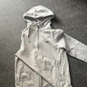 Vita tracksuit från Lacoste - En snygg vit dress från Lacoste med elastisk midja och dragsko. Byxorna har ribbade muddar vid bensluten och klassisk Lacoste-logga på benet. Perfekta för en chill och sportig look. Tillverkade i mjuk bomull för extra komfort.