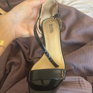 Svarta Michael Kors klackar  - Stilrena svarta sandaler från Michael Kors med smala remmar och öppen tå. Skorna har lackad finish, smal klack och guldfärgat spänne vid ankeln. Perfekta för dig som gillar en klassisk och elegant look.