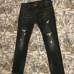 Svarta slim jeans från Crosshatch - Svarta jeans från Crosshatch i slim fit-modell med slitna detaljer och snygga slitningar framtill. Klassisk femficksdesign, kontrastsömmar och läderpatch bak i midjan. Jeansen har en mörk tvätt och är tillverkade i jeansmaterial.