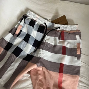 Rutig Burberry shorts med snörning - Snygga shorts från Burberry med klassiskt rutigt mönster i vitt, svart, beige, rosa och rött. De har elastisk midja med svart snörning och sidofickor. Perfekta för dig som vill sticka ut med en lyxig och trendig look.