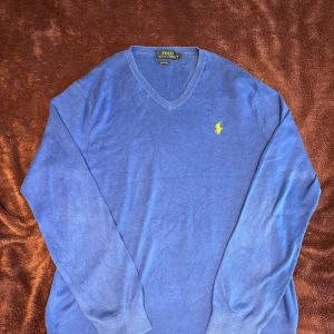 Blå tröja från Polo Ralph Lauren - Säljer en stilren blå tröja från Polo Ralph Lauren i pima bomull. Tröjan har en v-ringad design och det ikoniska gula logotypbroderiet på bröstet. Perfekt för en klassisk och avslappnad look. OBS! Etiketten på nacken sitter löst på ena änden.