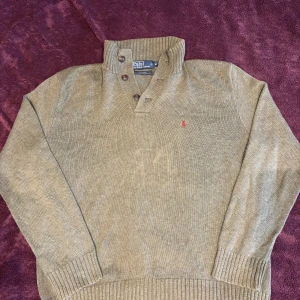 Stickad tröja från Polo Ralph Lauren - Säljer en stilren stickad tröja från Polo Ralph Lauren. Tröjan har en hög krage med knappar och det klassiska röda logotypbroderiet på bröstet. Perfekt för kyligare dagar.