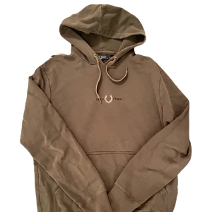 Grön hoodie från Fred Perry - Säljer en snygg grön hoodie från Fred Perry med klassisk logga på bröstet. Tröjan har en bekväm passform med långa ärmar och en praktisk huva med dragsko. Perfekt för en avslappnad stil.