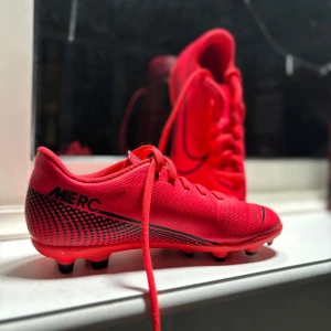Röda fotbollsskor från Nike - Snygga röda fotbollsskor från Nike med svarta detaljer och snörning. Perfekta för fotbollsplanen med sina dobbar för bra grepp. Skorna har en sportig design med Nike-logga på sidan.