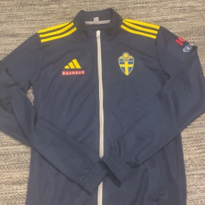 Blå träningsjacka från Adidas - Säljer en blå träningsjacka från Adidas med Sveriges landslagslogga. Jackan har gula ränder på axlarna och en dragkedja framtill hela vägen ner. Nyskick