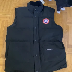 Canada goose väst - Säljer en svart dunväst från Canada Goose med deras ikoniska märke på bröstet. Västen har en hög krage och knappar framtill. Perfekt för kyliga dagar när du vill hålla värmen utan att kompromissa med stilen.