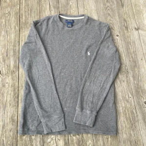Grå långärmad tröja från Polo Ralph Lauren - Säljer en stilren grå långärmad tröja från Polo Ralph Lauren. Tröjan är tillverkad i 100% bomull och har en broderad logotyp på bröstet. Perfekt för en avslappnad och snygg look.