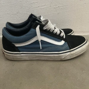 Svart och blå sneakers från Vans - Säljer ett par klassiska Vans sneakers i svart och blått med vita detaljer. Skorna har en ikonisk vit sidosöm och snörning framtill. Perfekta för en avslappnad stil.