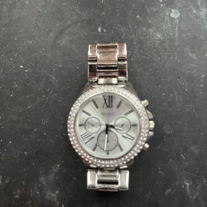 Silverfärgad klocka från Regal - Snygg silverfärgad klocka från Regal med glittrande stenar runt urtavlan. Klockan har romerska siffror och tre mindre urtavlor för extra funktioner. Perfekt accessoar för att lyfta din stil. Behövs byta batteri