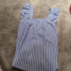 Blå- och vitrandig top - Charmig blå- och vitrandig top med volangdetaljer på axelbanden. Klänningen har en söt rosett framtill och är perfekt för en somrig look. Tillverkad i ett lätt och bekvämt material.