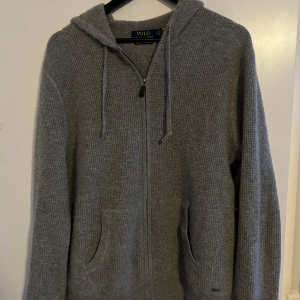 Grå stickad cashmere hoodie från Polo Ralph Lauren - Säljer en stilren grå stickad hoodie från Polo Ralph Lauren. Den har en dragkedja framtill och praktiska fickor. Perfekt för kyliga dagar med sin mjuka och bekväma passform. Huvan ger extra värme och stil. Passar både till vardags och avslappnade tillfällen.