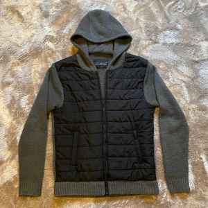 Svart och grå cardigan - Säljer en snygg svart och grå cardigan med huva från JOHN DEVIN. Säljer den då jag inte längre är i behov av den. Jackan har en quiltad framsida med dragkedja och stickade ärmar och rygg. Perfekt för kyliga dagar och ger en stilren look. Priset kan såklart diskuteras men det är bara hör av er om ni har några frågor! 