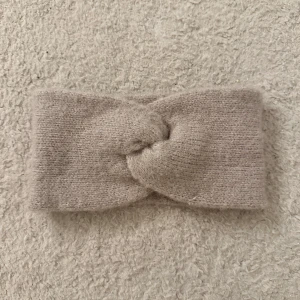 Beige stickat huvudvärmare - Säljer en mysig, beige, stickad huvudvärmare. Perfekt för att hålla värmen och stilen under kyliga dagar. Huvudvärmaren har en enkel och stilren design med en twist framtill. Aldrig använd då den inte riktigt passar min stil💞
