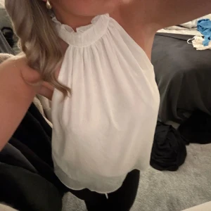 Vit ärmlös halterneck-top - Säljer en stilren vit halterneck-top med hög krage och ärmlös design. Toppen har en lös passform och knäpps i nacken. Perfekt för en elegant look.