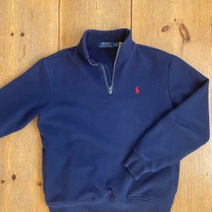  Polo Ralph Lauren half zip - Säljer en stilren mörkblå tröja från Polo Ralph Lauren med halv dragkedja och röd broderad logotyp på bröstet. Perfekt för en avslappnad och snygg look. Tröjan har långa ärmar och ribbade muddar.