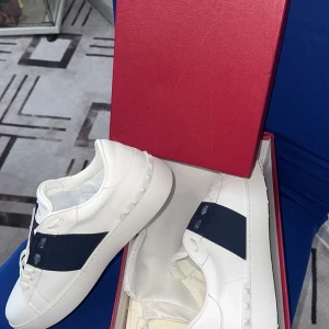 Valentinos garavani  - Snygga vita sneakers från Valentino med en bred marinblå rem över ovansidan. Skorna har en stilren design med diskreta nitar på sidorna och en bekväm sula. Perfekta för en trendig look. Jag valde sälja dem för de var alldeles för små för mig. 