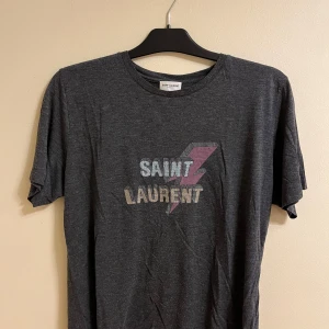 Grå t-shirt från Saint Laurent - Snygg grå t-shirt från Saint Laurent med ett coolt blixtmotiv och texten 'SAINT LAURENT' i metallicfärger på framsidan. Perfekt för en avslappnad stil med en touch av lyx.