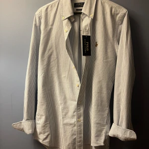 Randig skjorta från Ralph Lauren - Snygg och klassisk skjorta från Ralph Lauren i blått och vitt randigt mönster. Skjortan har långa ärmar och knappar framtill samt en broderad logotyp på bröstet. Perfekt för en stilren look! Skjortan är HELT NY bara testad en gång och jag säljer denna pga att den är för stor för mig! pris kan diskuteras vid en snabb affär! 