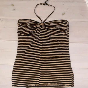 Randig tubtopp från H&M - Säljer en snygg randig tubtopp från H&M i svart och beige. Toppen har en halterneck-design med smala band och en åtsittande passform. Perfekt för varma sommardagar eller en kväll ute. 🖤