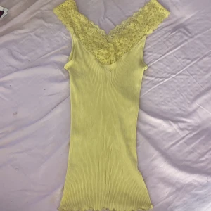 Gul ribbad topp med spetsdetaljer - Säljer en söt gul ribbad topp med vacker spetsdetalj vid halsringningen. Toppen har en v-ringad design och är perfekt för varmare dagar. Den är både stilren och bekväm. Den sitter tajt på och är lite längre topp. Jag har storlek XS i linnen och denna sitter perfekt även om de är storlek M