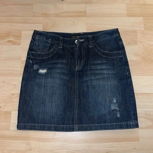Jeanskjol mörkblå med slitningar - Snygg jeanskjol i mörk denim med slitningar framtill. Kjolen har klassiska femficksdetaljer och en knappstängning. Perfekt för en avslappnad stil.