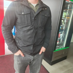 Svart jacka från jj lindberg - Snygg svart jacka från j lindberg med knappar och dragkedja. Jackan har långa ärmar och flera praktiska fickor framtill. Perfekt för en stilren look. 