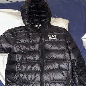 Svart pufferjacka från Emporio Armani - Säljer en svart pufferjacka från Emporio Armani med huva och dragkedja. Jackan har ett stilrent quiltat mönster och EA7-logga på bröstet. Perfekt för kyliga dagar. 🖤(Den har blivit tejpad med svart tejp på en jätte liten del på armen. Inget man tänker på)