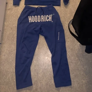 Blå mjukisbyxor från Hoodrich - Snygga blå mjukisbyxor från Hoodrich med stort tryck på framsidan. Byxorna har en praktisk ficka på sidan och snörning i midjan för bästa passform. Perfekta för en avslappnad stil.