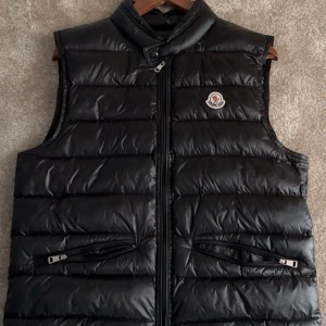 Svart dunväst från Moncler - Snygg svart dunväst från Moncler med dragkedja och två fickor framtill. Perfekt för kyligare dagar och ger en stilren look. Västen har en diskret logga på bröstet.