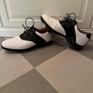Svartvita golfskor FootJoy  - Snygga svartvita golfskor från FootJoy med snörning och spikar på sulan för extra grepp. Perfekta för golfbanan med en stilren design i svart och vitt. Storlek 43 