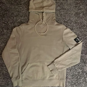 Beige hoodie från Calvin Klein Jeans - Säljer en stilren beige hoodie från Calvin Klein Jeans. Den har en klassisk design med en justerbar huva och en praktisk magficka. Perfekt för en avslappnad look.             Nypris:1400
