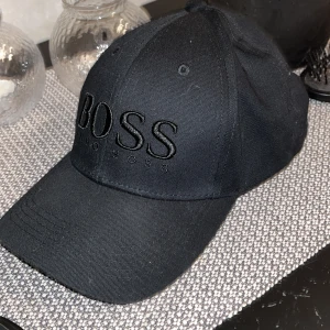 Svart keps från Hugo Boss - Snygg svart keps från Hugo Boss med broderad logotyp framtill. Kepsen har en justerbar rem baktill för perfekt passform. Perfekt för en stilren look.