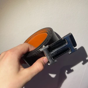 Svart bälte med silverfärgat spänne - Snyggt svart bälte med ett iögonfallande silverfärgat H-format spänne. Insidan av bältet är i en kontrasterande orange färg. Perfekt för att ge en stilren touch till din outfit.