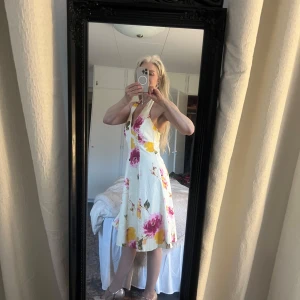 Blommig halterneckklänning med volangdetaljer - Charmig vit klänning med stora blommor i rosa och gult. Den har en v-ringad hals och volangdetaljer vid fållen. Perfekt för sommarens festligheter!