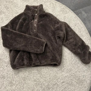 Brun fleece tröja med knappar - Mysig brun fleece tröja med hög krage och knappar framtill. Perfekt för kyliga dagar med sin mjuka och varma känsla. Tröjan har långa ärmar och en avslappnad passform.