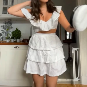 Vit klänning med volanger - Charmig vit klänning med volanger och kort längd. Den har en v-ringad topp med volangdetaljer och är ärmlös. Perfekt för studenten!!