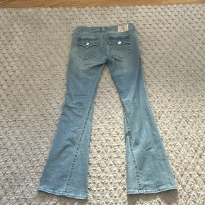 Blå jeans från True Religion - Säljer ett par blå jeans från True Religion med låg midja och utsvängda ben. Modellen heter Joey Low Rise Flare. Jeansen har en klassisk femficksdesign och knappar med märkets logotyp. Använd endast 1 gång då de va förstora.