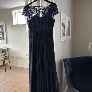 Mörkblå långklänning från Vila - Elegant mörkblå långklänning från Vila med spetsdetaljer och knappar baktill. Klänningen har korta ärmar och en vacker, genombruten spetsdesign upptill. Perfekt för en formell tillställning.