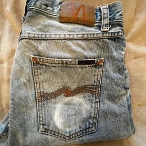 Blå jeans från Nudie Jeans - Säljer ett par ljusblå jeans från Nudie Jeans, modell Steady Eddie II. De är tillverkade av 100% ekologisk bomull och har en klassisk femficksdesign med orange sömmar. Perfekta för en avslappnad stil.