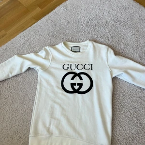 Vit tröja från Gucci - Säljer en stilren vit tröja från Gucci med det ikoniska logotyptrycket i svart på framsidan. Tröjan har långa ärmar och en klassisk rund halsringning. Perfekt för en avslappnad men lyxig look.
