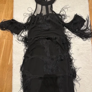 Svart klänning med fransar - Snygg svart klänning med unika fransdetaljer och offshoulder-design. Perfekt för en kväll ute eller en speciell tillställning. Klänningen har en elegant och modern look med en tight passform.