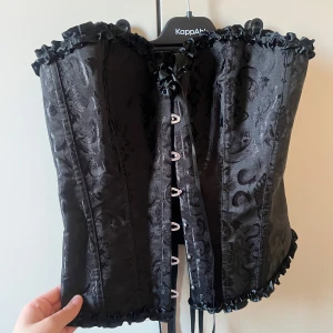 Svart korsett med brokadmönster - Elegant svart korsett med vackert brokadmönster som aldrig kommit till användning!🩷 Den har snörning framtill och volangdetaljer längs kanterna. Perfekt för en stilfull look. 