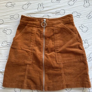 Brun/orange manchesterkjol från New Yorker - Säljer en snygg brun/orange manchesterkjol från New Yorker i storlek XS. Kjolen har en dragkedja framtill med en rund ringdetalj och praktiska fickor. Perfekt för en trendig look!