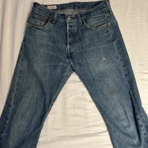 Levis 501 jeans i blå denim - Klassiska Levis 501 jeans i blå denim. De har en rak passform och knappar i gylfen. Perfekta för en avslappnad stil med fem fickor och den ikoniska Levis-lappen bak. Passar till allt och är ett måste i garderoben.